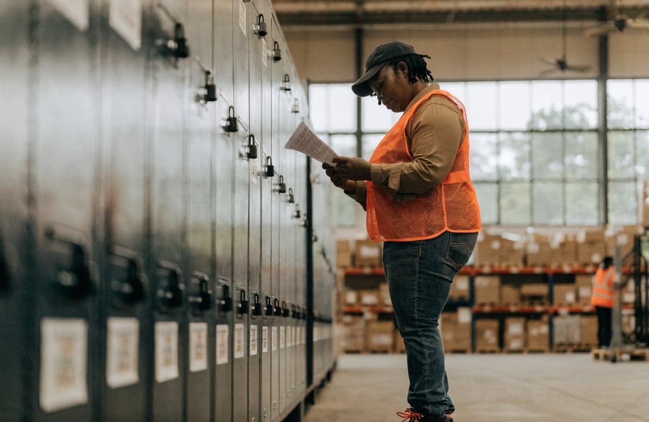 Warehouse worker in a safety vest reading a notice beside a row of lockers