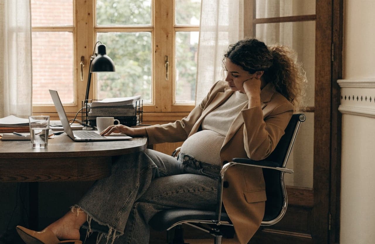 Pregnant employee pausing while working on a laptop at an office desk